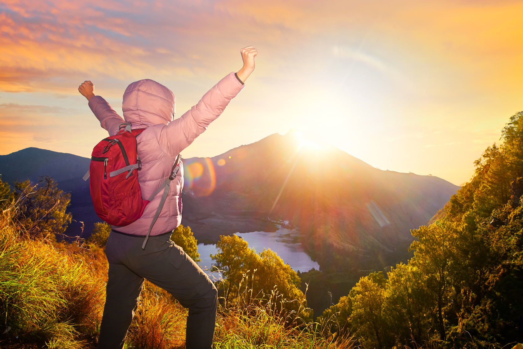 happy traveler with backpack standing early morning on a rock with raised hands looking to a volcano Rinjani, island Lombok. Indonesia.